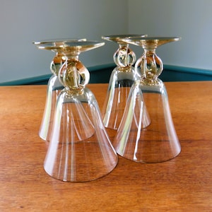 May include: Four clear glass goblets with a gold-colored stem and base are arranged on a wooden surface. The goblets are upside down, showcasing their bell-shaped bowls and decorative stems. The background is a muted blue and green.