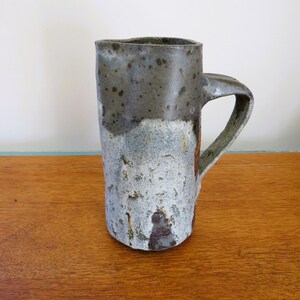 Puede incluir: Una taza alta de cerámica hecha a mano con una superficie texturizada única. La taza presenta una parte superior gris oscuro, que pasa a una base gris claro y blanca. Tiene un asa resistente y se exhibe sobre una superficie de madera. La taza mide unos 15 cm de alto.