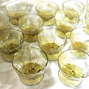 May include: A collection of fourteen amber-colored glass dessert bowls. Each bowl features a textured, irregular surface with a wide, rounded rim and a short, sturdy pedestal base. The glass has a translucent, golden-yellow hue throughout.