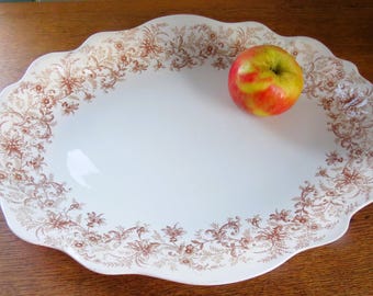 May include: An oval, white serving platter with a scalloped edge and a brown floral pattern. A red and yellow apple rests on the platter. The platter is on a wooden surface.