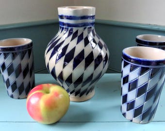 May include: A still life featuring a ceramic vase and three matching tumblers, all with a blue and white diamond pattern. A red and yellow apple sits in front of the objects. The objects are on a light blue surface.