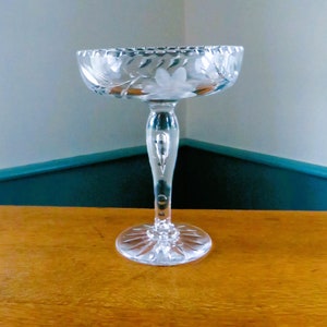 May include: A clear glass bowl with a decorative, scalloped rim and a floral pattern. It has a long, slender stem and a circular base. The glass is cut with a pattern. It is likely used for serving desserts or cocktails.