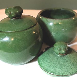 May include: Three-piece green ceramic set. Includes a lidded sugar bowl with a frog-shaped handle, an open bowl, and a lid with a frog-shaped handle. The set has a glossy finish and a rounded shape.