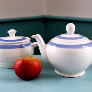 May include: White ceramic teapot and lidded sugar bowl with blue stripes. A red apple sits in front of the teapot and bowl. The teapot has a curved spout and handle. The bowl has two handles.