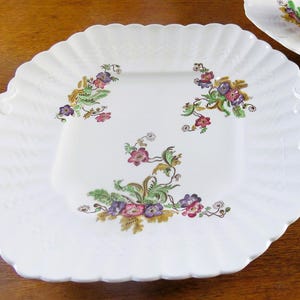 May include: White, scalloped-edge serving platter with floral designs in purple, pink, green, and yellow. The platter has two handles and a square center. A second platter is partially visible in the background.