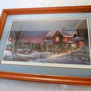 May include: Framed print of a winter scene with a snow-covered road leading to a warmly lit house. The print is set within a light blue mat and a brown wooden frame, creating a cozy, inviting aesthetic.