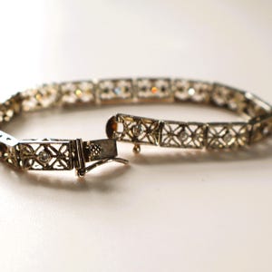 May include: An open, ornate bracelet with a silver-colored metal finish. The bracelet features a series of square links with a delicate, openwork design. Small, clear gemstones are set within the links. The clasp is open, revealing the intricate details.
