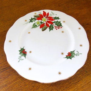 May include: White ceramic plate with a scalloped edge, decorated with a poinsettia, holly, and gold starbursts. The plate has a gold rim and is suitable for holiday dining.