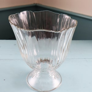 Puede incluir: Un jarrón de cristal transparente con base y borde festoneado. Presenta un diseño acanalado vertical y una base redondeada. El jarrón es ideal para flores o como elemento decorativo. La luz pasa a través del cristal.