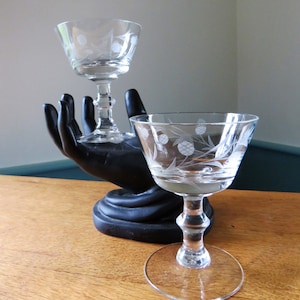 Puede incluir: Dos copas de cóctel de cristal transparente con diseños de cardos grabados. Una copa está en posición vertical, la otra la sostiene una escultura negra con forma de mano. Las copas están sobre una superficie de madera, con un fondo neutro.