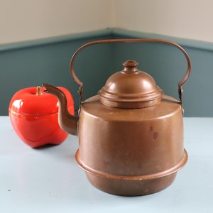 May include: A copper tea kettle with a rounded body, a curved spout, and a matching lid with a small knob handle. The kettle features a tall, arched metal handle attached to the sides. Beside the kettle sits a bright red, apple-shaped container.