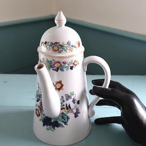 Op de afbeelding: Witte keramische koffiepot met deksel, versierd met een bloemenpatroon in rood, blauw, geel en groen. Het heeft een gebogen handvat en een tuit. Het deksel is bekroond met een goudkleurige knop.