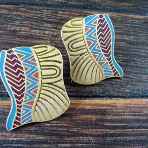 Puede incluir: Dos pendientes dorados con un diseño en forma de pluma. Cada pendiente presenta un patrón geométrico azul, morado y naranja sobre una base dorada. Los pendientes están colocados sobre un fondo de vetas de madera oscura.