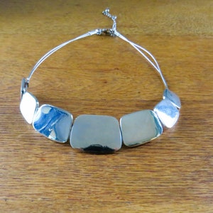 Può includere: Una collana argentata con una serie di piastre quadrate in metallo lucido. Le piastre sono collegate da sottili fili d'argento flessibili, creando un design moderno. La collana è esposta su una superficie di legno.