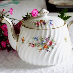 May include: A white porcelain teapot featuring a ribbed, swirling texture and a delicate floral pattern in shades of pink, blue, and orange. The teapot includes a matching lid with a gold-trimmed handle and spout, accented by gold detailing.
