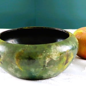 Peut inclure: Un bol vert et noir fait à la main avec une finition texturée. À côté du bol se trouve une pomme rouge et jaune. Le bol a une forme arrondie et un intérieur sombre. L'arrière-plan est de couleur sarcelle unie.