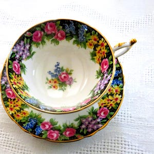Op de afbeelding: Bloemen theekop en schotel set. De witte theekop en schotel zijn versierd met een zwarte rand en een kleurrijk bloemenpatroon van roze rozen, gele bloemen en blauwe en paarse bloesems. De rand en het handvat zijn goudkleurig.