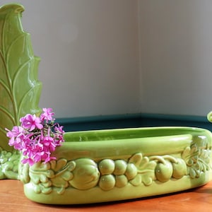 May include: A set of light green ceramic vessels featuring a tall leaf-shaped vase and an oval planter with embossed fruit and leaf motifs. A small cluster of bright pink phlox flowers rests inside the oval planter, adding a vibrant color contrast.