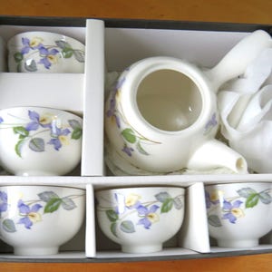 Könnte beinhalten: Ein Teeservice in einer Schachtel mit einer Teekanne mit seitlichem Griff und fünf passenden Teetassen. Die cremeweißen Keramikstücke zeigen ein zartes Blumenmuster mit violetten und gelben Blütenblättern sowie grünen Blättern.