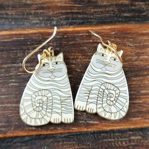 May include: A pair of dangle earrings featuring a cat design with cream and light gray wavy stripes. The cat figures are crafted with gold-toned metal outlines and matching gold-toned ear wires for hanging. Each cat has a curled tail and whiskers.
