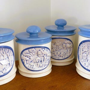 May include: Four ceramic canisters with blue lids and oval relief scenes. The canisters are off-white with blue accents. Each canister features a different rural landscape scene in a blue oval frame.