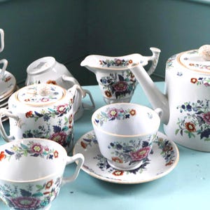 Könnte beinhalten: Ein weißes Porzellan-Teeservice mit einem bunten Blumenmuster in Rosa, Blau und Orange. Das Set besteht aus einer Teekanne mit Deckel, einem Milchkännchen, einer Zuckerdose mit Deckel sowie mehreren Teetassen mit passenden Untertassen und Goldrand.