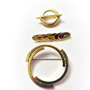May include: Three gold-toned brooches are displayed against a white background. The brooches include a circular design with a horizontal bar, a series of connected ovals, and a large open circle with cut-out sections. The jewelry pieces are likely vintage or retro.