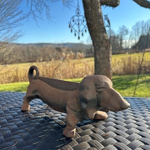 May include: A brown cast iron dachshund dog sculpture. The dog is in a resting position with its head down and tail curled up. The sculpture is sitting on a black woven surface, with a natural outdoor background.