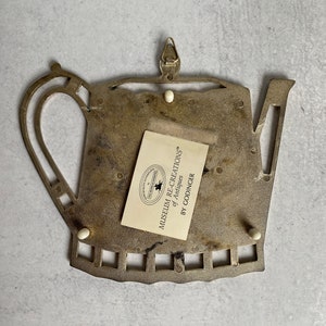 May include: A metal teapot-shaped trivet with a decorative design. The trivet has a label that reads "Museum Recreations of Antiques by Godinger".