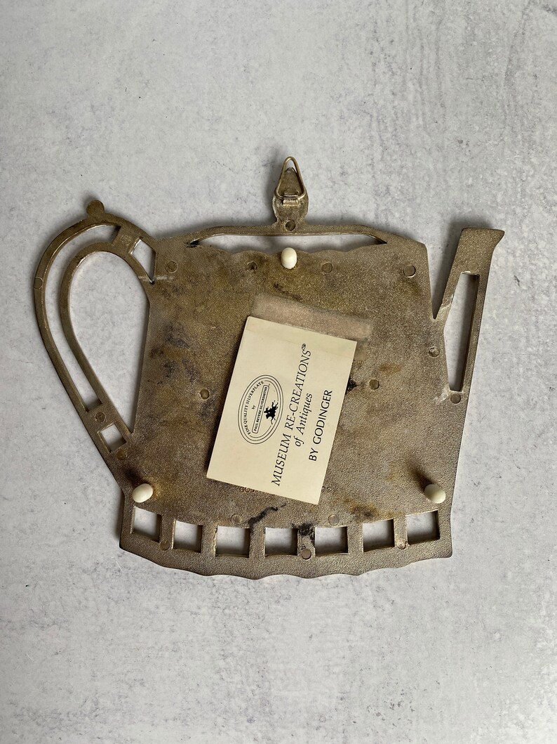 May include: A metal teapot-shaped trivet with a decorative design. The trivet has a label that reads "Museum Recreations of Antiques by Godinger".