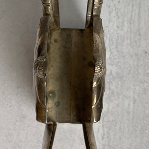 May include: Silver napkin holder with a decorative design. The holder is shaped like a pair of crossed arms with a rectangular opening in the center.