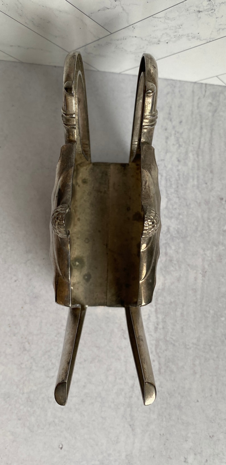 May include: Silver napkin holder with a decorative design. The holder is shaped like a pair of crossed arms with a rectangular opening in the center.