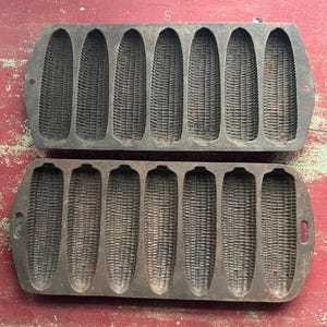 May include: Two antique cast iron corn-shaped bread molds. Each mold has seven corn cob impressions. The molds are dark gray and show signs of wear, suggesting they are vintage kitchenware. The molds are on a red surface.
