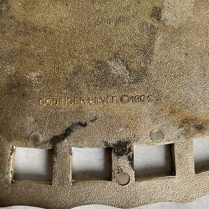 May include: Close-up of a gold-colored metal plate with a scalloped edge. The plate has a textured surface and several square cutouts. The text "SPRINGFIELD SILVER 1992" is engraved on the plate.