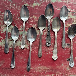 May include: An assortment of antique silver spoons, each with unique designs, arranged on a distressed red wooden surface. The spoons vary in size and style, showcasing intricate patterns and ornate handles. A vintage collection.