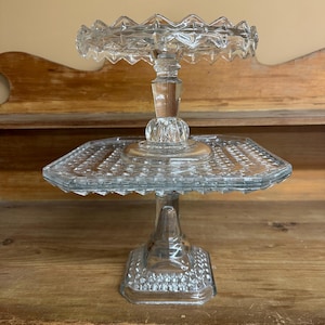 May include: A two-tiered clear glass serving stand featuring a square base and a square middle plate with a textured diamond pattern. The top tier is a circular plate with a scalloped, jagged edge, supported by a central glass pedestal.