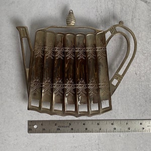 May include: Silver-toned metal teapot-shaped utensil holder with an ornate design. The holder has six slots for utensils and a decorative knob on the top.