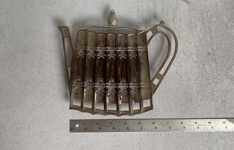 May include: Silver-toned metal teapot-shaped utensil holder with an ornate design. The holder has six slots for utensils and a decorative knob on the top.