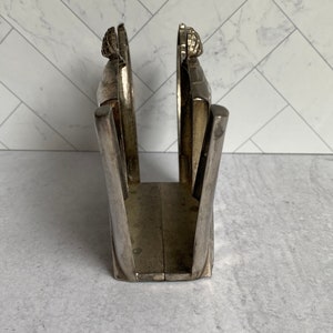 May include: Silver napkin holder with a decorative design. The holder is made of metal and has a rectangular shape.