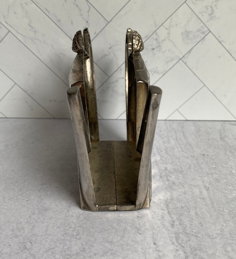 May include: Silver napkin holder with a decorative design. The holder is made of metal and has a rectangular shape.