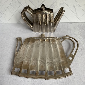 May include: Silver teapot stand with an ornate design. The stand is shaped like a teapot and has a scalloped edge. The stand is made of metal and has a shiny finish.