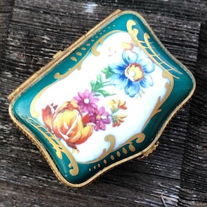 May include: A small, hinged, green and gold trinket box with a white porcelain lid featuring a hand-painted floral design. The flowers are in shades of blue, pink, yellow, and orange.