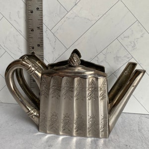 May include: Silver teapot napkin holder with a decorative design. The holder is shaped like a teapot with a spout, handle, and lid.