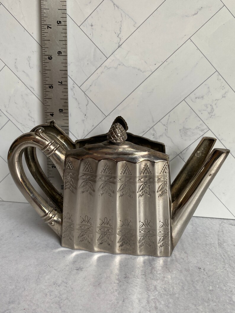 May include: Silver teapot napkin holder with a decorative design. The holder is shaped like a teapot with a spout, handle, and lid.