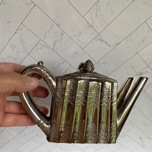 May include: A silver teapot with a decorative design and a pine cone finial. The teapot has a curved spout and a handle.