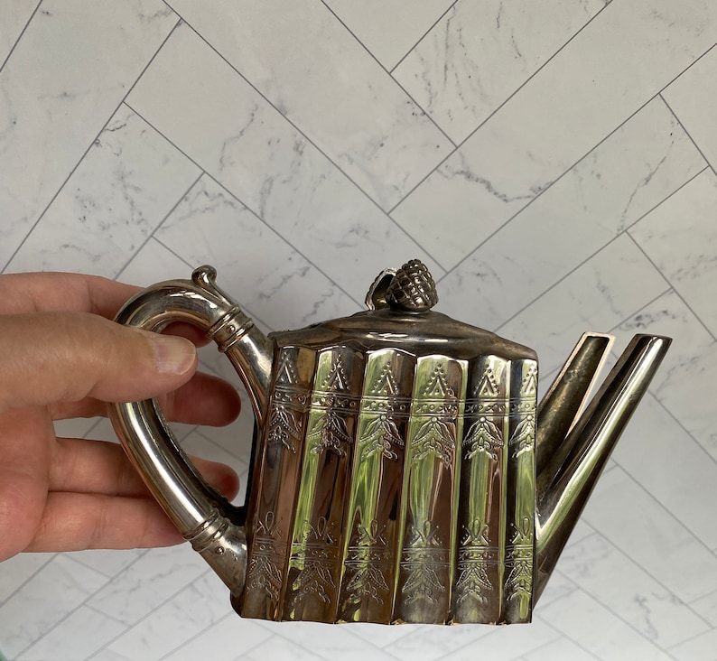May include: A silver teapot with a decorative design and a pine cone finial. The teapot has a curved spout and a handle.