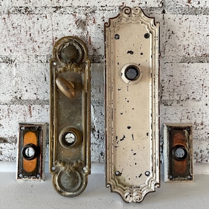 May include: An assortment of antique door hardware, including two ornate door plates and two rectangular doorbell buttons. The door plates are aged metal with intricate designs, and the doorbell buttons have a dark center.