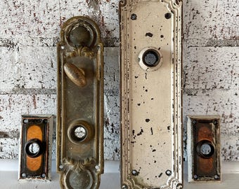 Pode incluir: Uma variedade de ferragens de portas antigas, incluindo duas placas de porta ornamentadas e dois botões de campainha retangulares. As placas de porta são de metal envelhecido com designs intrincados, e os botões de campainha têm um centro escuro.