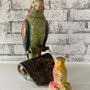 May include: A vintage, hand-painted parrot figurine with red, green, and blue coloring, perched on a wooden stand. A small owl figurine and a rolled-up scroll are also present. The background is a white brick wall.