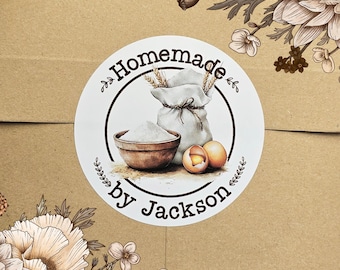 May include: White round sticker with brown text that reads "Homemade by Jackson". The sticker features a drawing of a bag of flour, a bowl of flour, and two eggs.